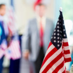 A person is holding an American flag while a politician makes a speech during political rally. Focus is on the American flag in the foreground.