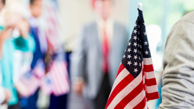 A person is holding an American flag while a politician makes a speech during political rally. Focus is on the American flag in the foreground.